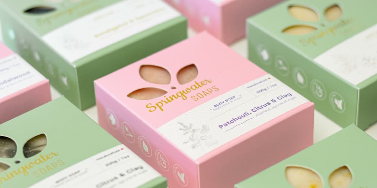 Cajas de jabón artesanal en rosa y verde alineadas sobre una superficie clara